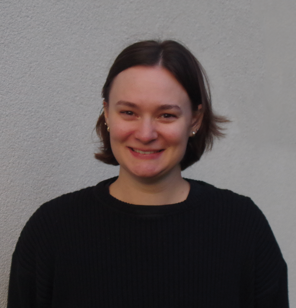 Morgan King is looking at the camera smiling shoulders and above, there is a textured white background, she is wearing a black sweater and is a white woman with brown hair to her chin and brown eyes