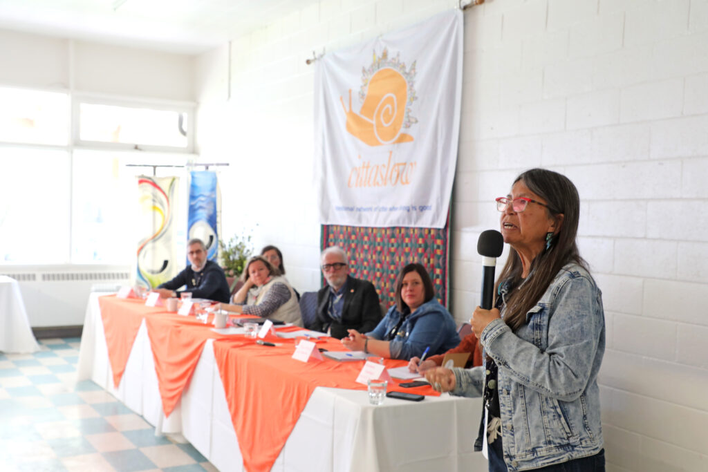 Pauline Terbasket, Syilx Okanagan woman, speaking with microphone to the right and the panel of experts to the left seated at a table under the cittaslow sign