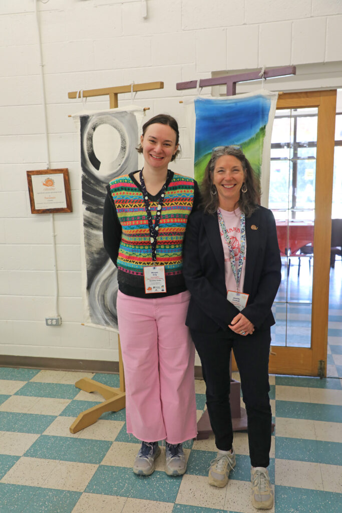 Morgan and Jennifer Cockrall smiling for the camera, Morgan is wearing a colourful vest and pink pants. Jennifer is wearing a pink shirt, black blazer, and black pants