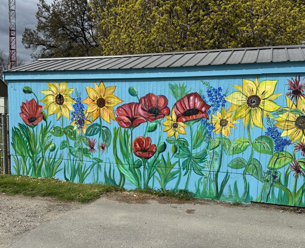 A mural on the side of a building, with the background a light blue and large flowers covering it. Poppies, sunflowers, and lilacs, with many green leaves.